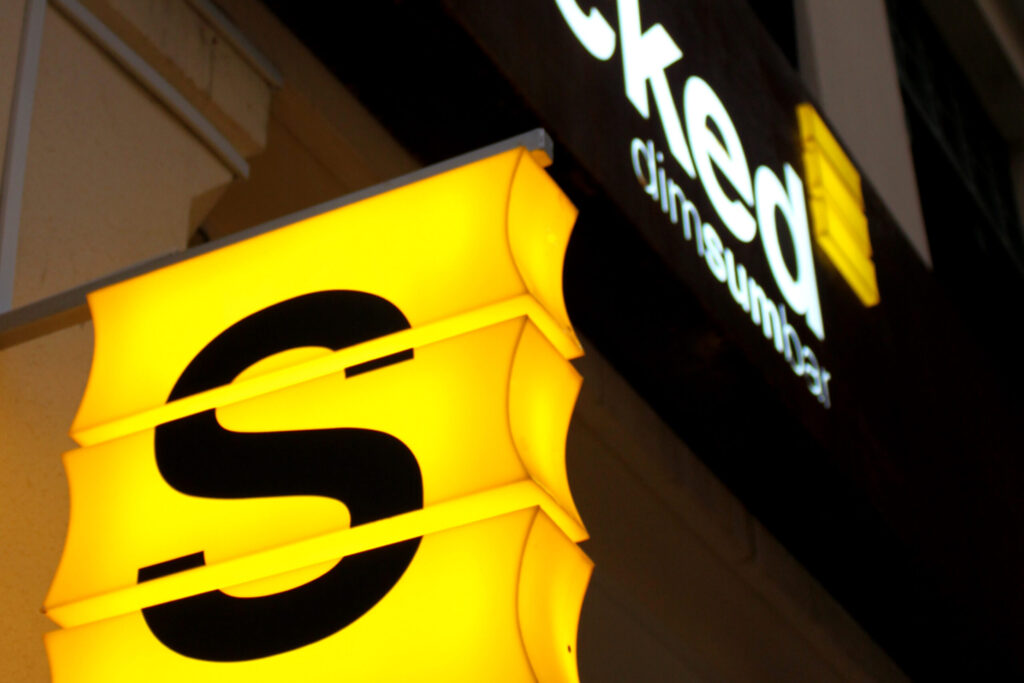 Bright yellow sign with a large black letter 'S' in the foreground, with a dimly lit sign reading 'dim sum bar' visible in the background.